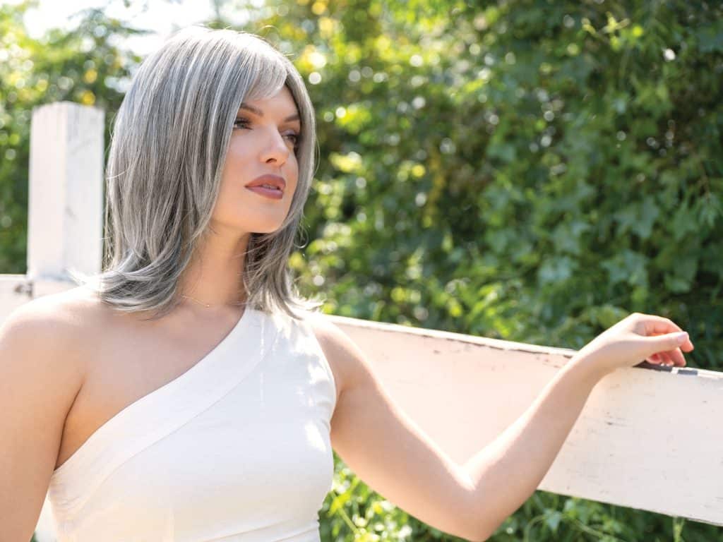 Woman with silver-gray wig outdoors, natural sunlight, lush green background.